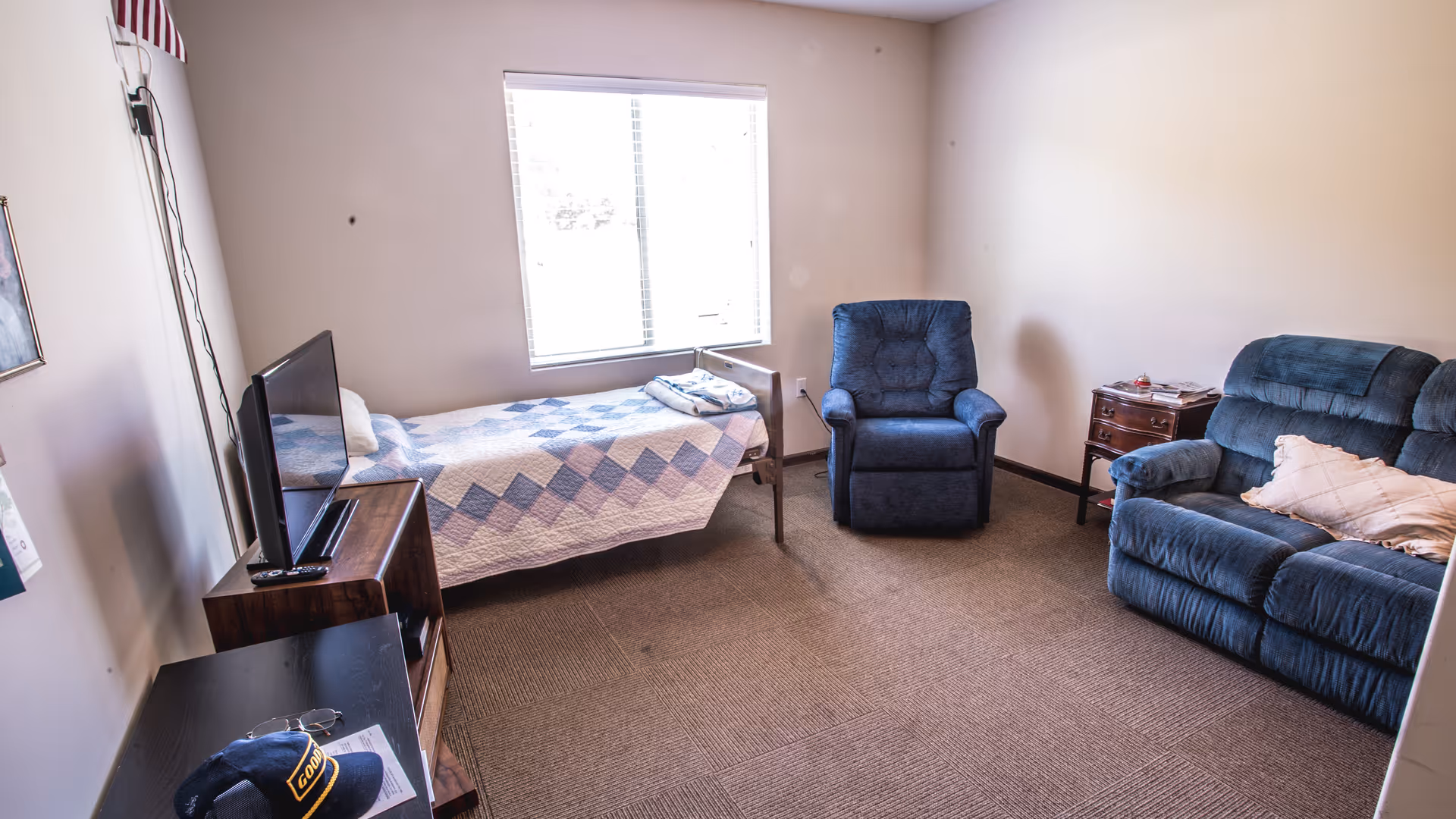 A small room with a single bed covered with a blue and white quilt, a blue armchair, a blue loveseat with a pillow, a wooden nightstand with items on it, a TV on a wooden stand, and a window letting in natural light.