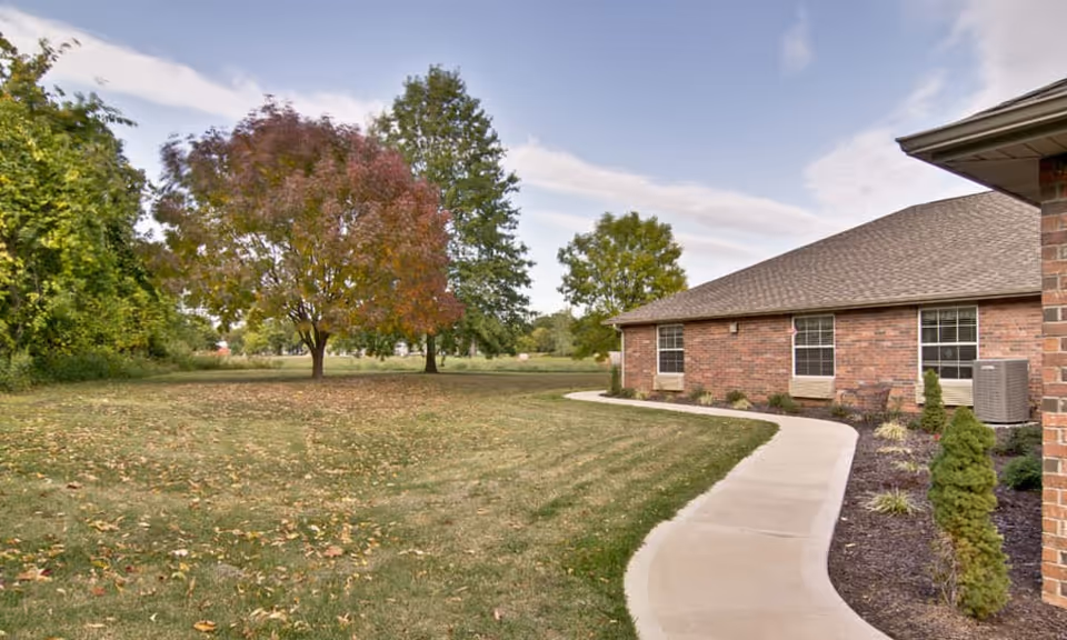 A curved concrete walkway beside a brick building with windows, surrounded by a landscaped area with small shrubs and trees. In the background, there is a grassy lawn with scattered fallen leaves and several trees with green and autumn-colored foliage under a partly cloudy sky.