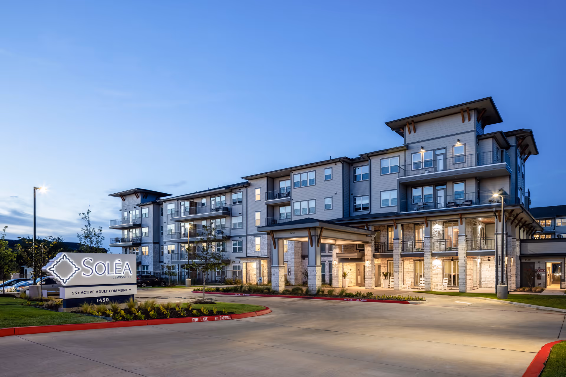 Exterior view of Solea Lewisville, a modern multi-story building with balconies and well-lit entrance, surrounded by landscaped greenery and a parking area at dusk.