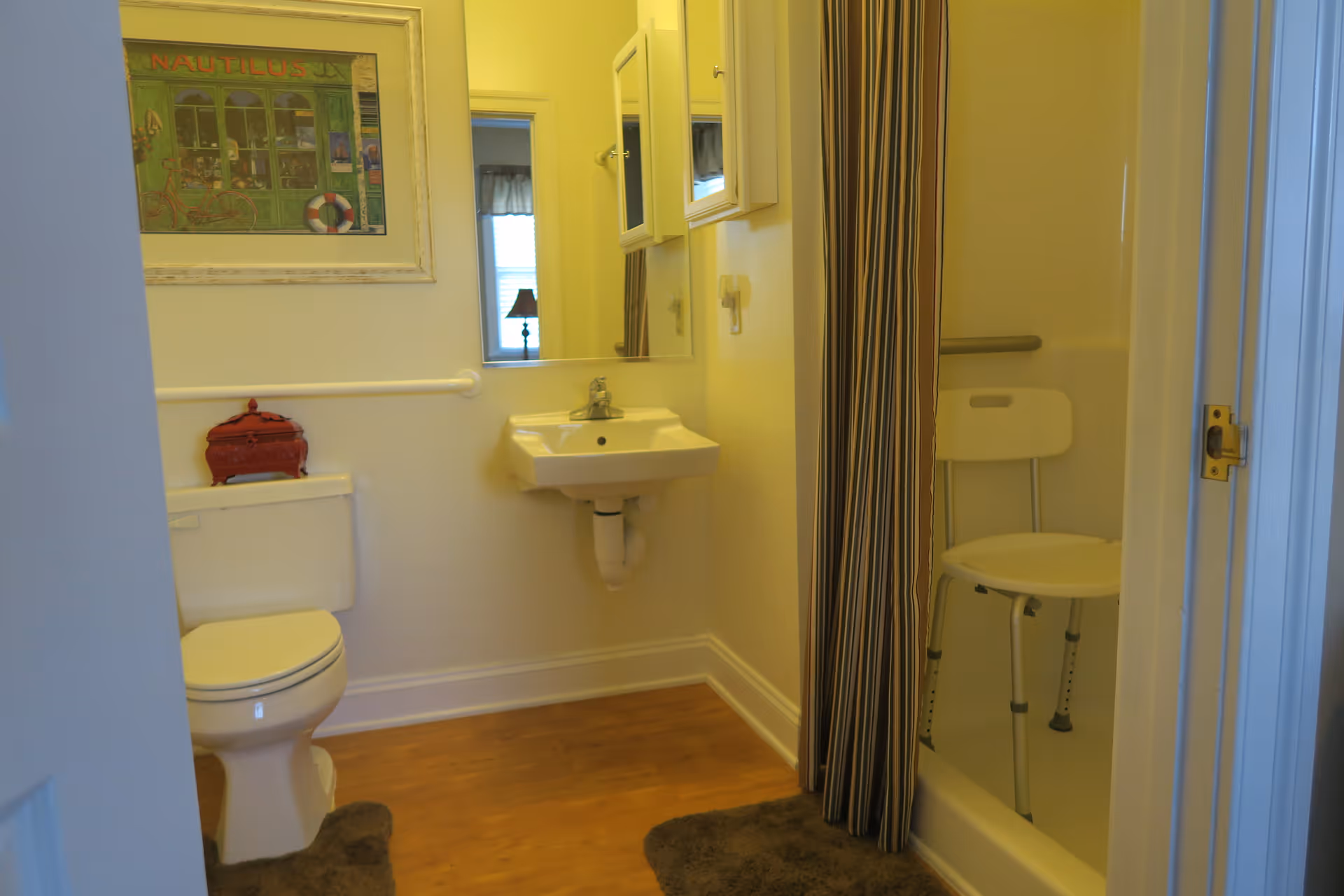 Small bathroom with a toilet, wall-mounted sink and mirror, and a shower containing a folding shower chair behind a striped curtain.