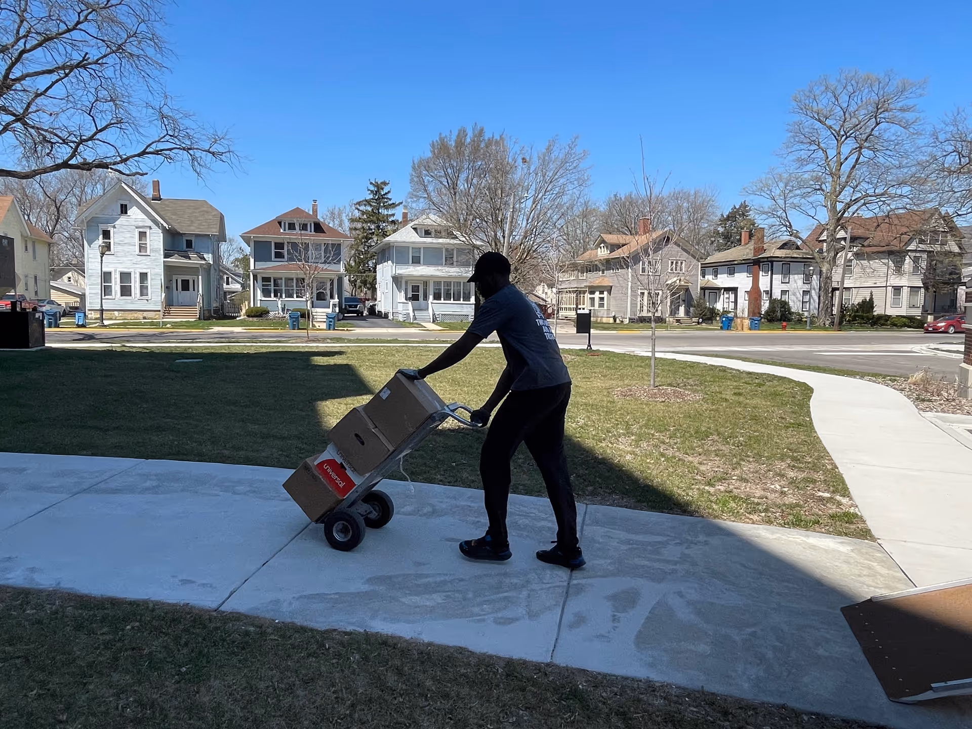 A person pushing a hand truck loaded with cardboard boxes on a sidewalk outside, with residential houses and trees in the background under a clear blue sky.