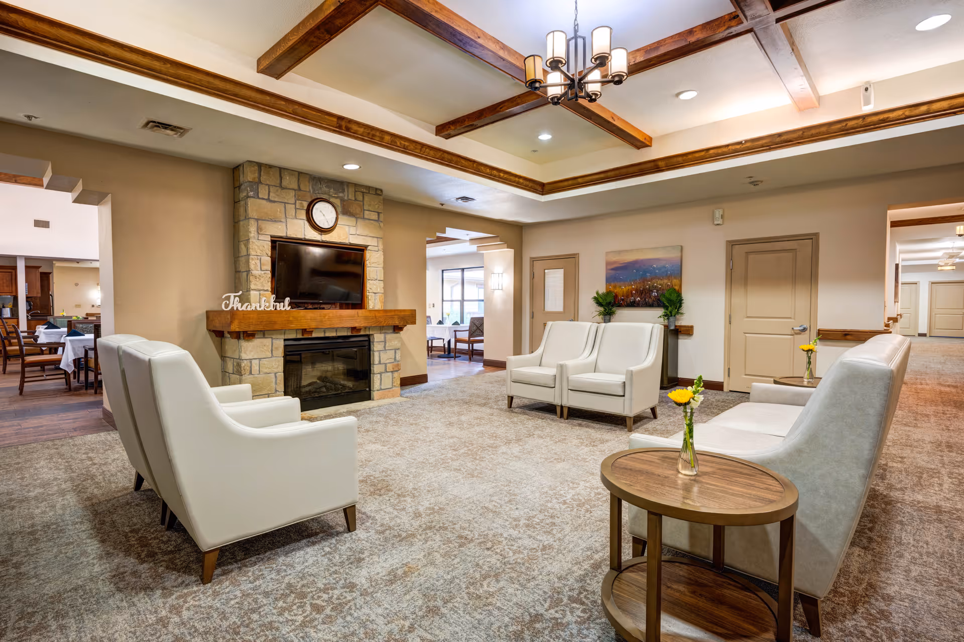 A cozy living room area in Oak Hills Terrace Memory Care featuring white armchairs and sofas arranged around a stone fireplace with a mounted TV above it. The room has a beige carpet, wooden ceiling beams, a chandelier, and small tables with yellow flowers in vases. Adjacent dining areas are visible through open doorways.