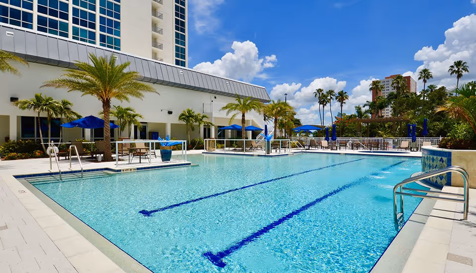 Outdoor swimming pool area at The Palms of Fort Myers with clear blue water, palm trees, blue umbrellas, lounge chairs, and a bright sunny sky with scattered clouds.