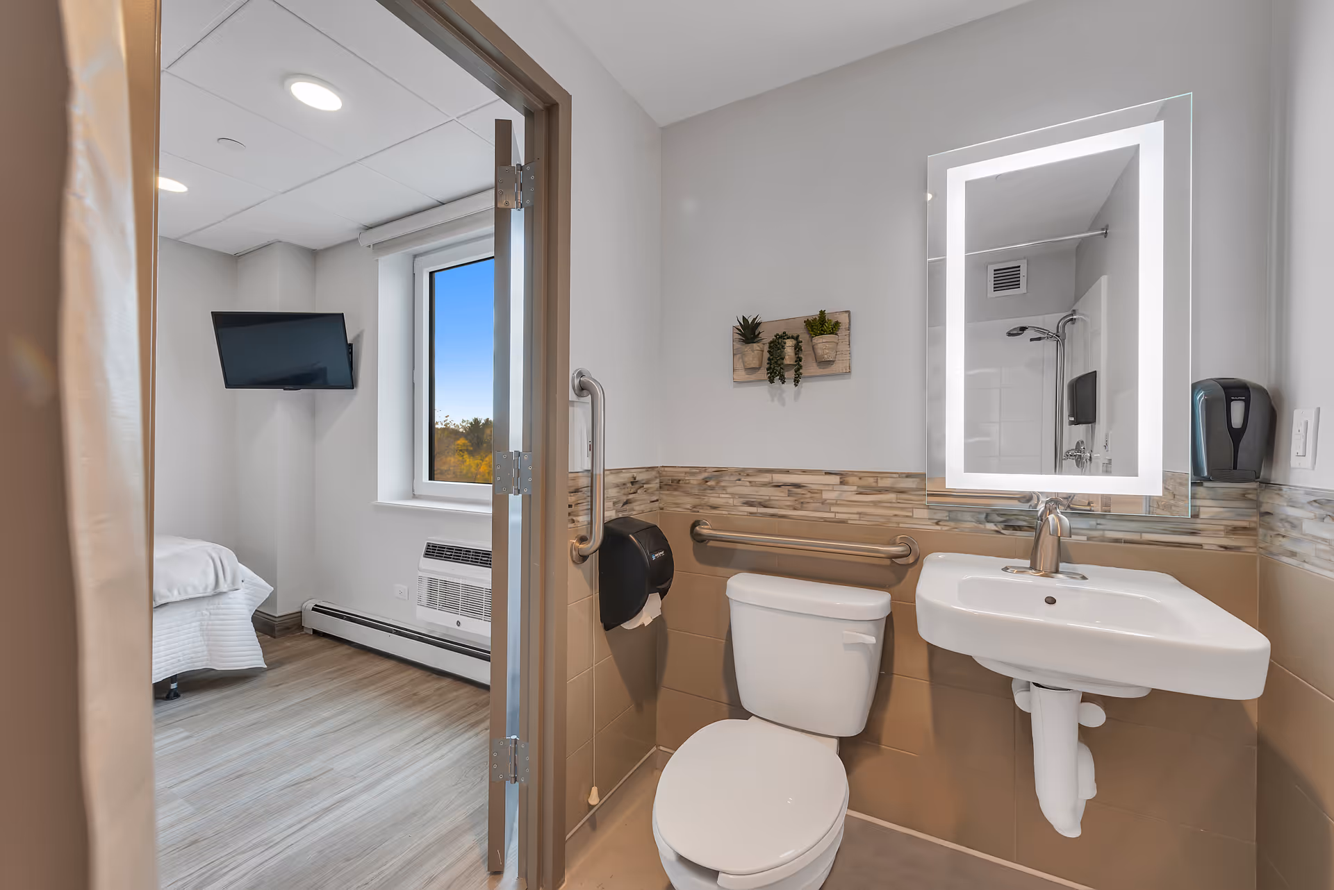 A clean and modern bathroom with a white toilet, a wall-mounted sink, and a rectangular mirror with built-in lighting. The walls have a beige tile halfway up with a decorative tile border. There are grab bars near the toilet and sink for accessibility. A small wall decoration with three potted plants hangs above the toilet. The bathroom door is open, showing a glimpse of a bedroom with a bed, a window, and a wall-mounted TV.