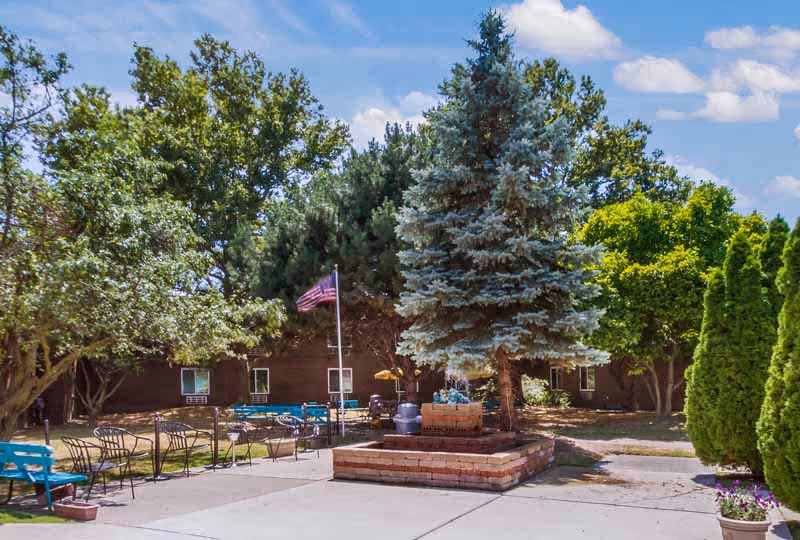 Outdoor courtyard area with a large evergreen tree planted in a raised brick planter in the center. Surrounding the courtyard are several metal chairs and benches, some painted blue. There is an American flag on a flagpole near the tree. The background shows a brown building with windows and various green trees under a partly cloudy blue sky.