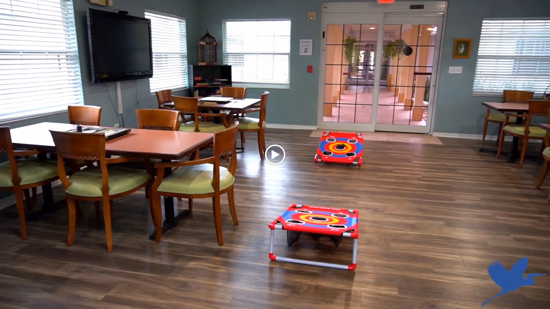A spacious room with wooden floors featuring several tables and chairs arranged for seating. Two red and blue bean bag toss game boards are set up on the floor. The room has large windows with blinds, a wall-mounted TV, and a glass door leading to an outdoor corridor.
