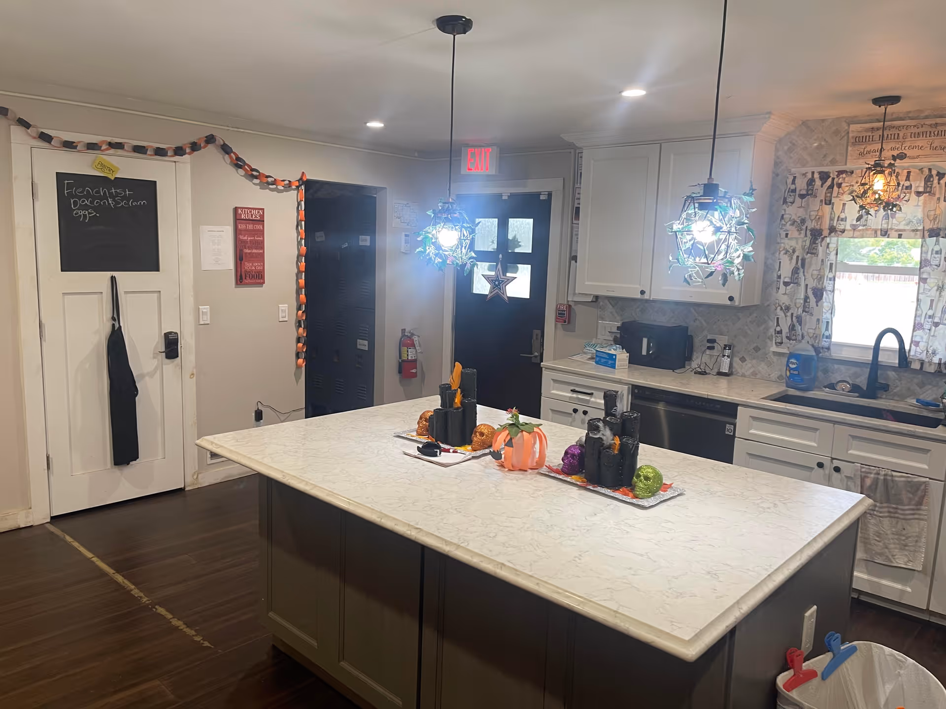 Interior view of a kitchen with a large marble island countertop decorated with Halloween-themed items including black candles, a pumpkin, and colorful skulls. The kitchen has white cabinets, a black sink, and a window with patterned curtains. Two pendant lights hang above the island. A door with a black apron hanging on it and a chalkboard with a breakfast menu written on it is visible on the left side. The floor is dark wood, and there is an exit door in the background.