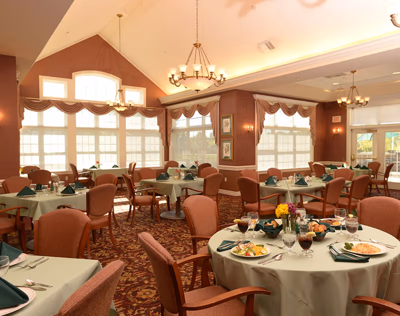 Dining room with round tables set for a meal, upholstered chairs, chandeliers, and large arched windows.