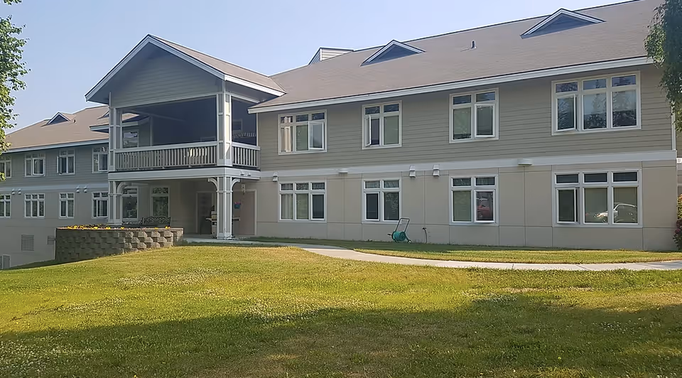 Two-story senior living facility building with a covered balcony entrance, rows of windows, and a grassy lawn and walkway in front.