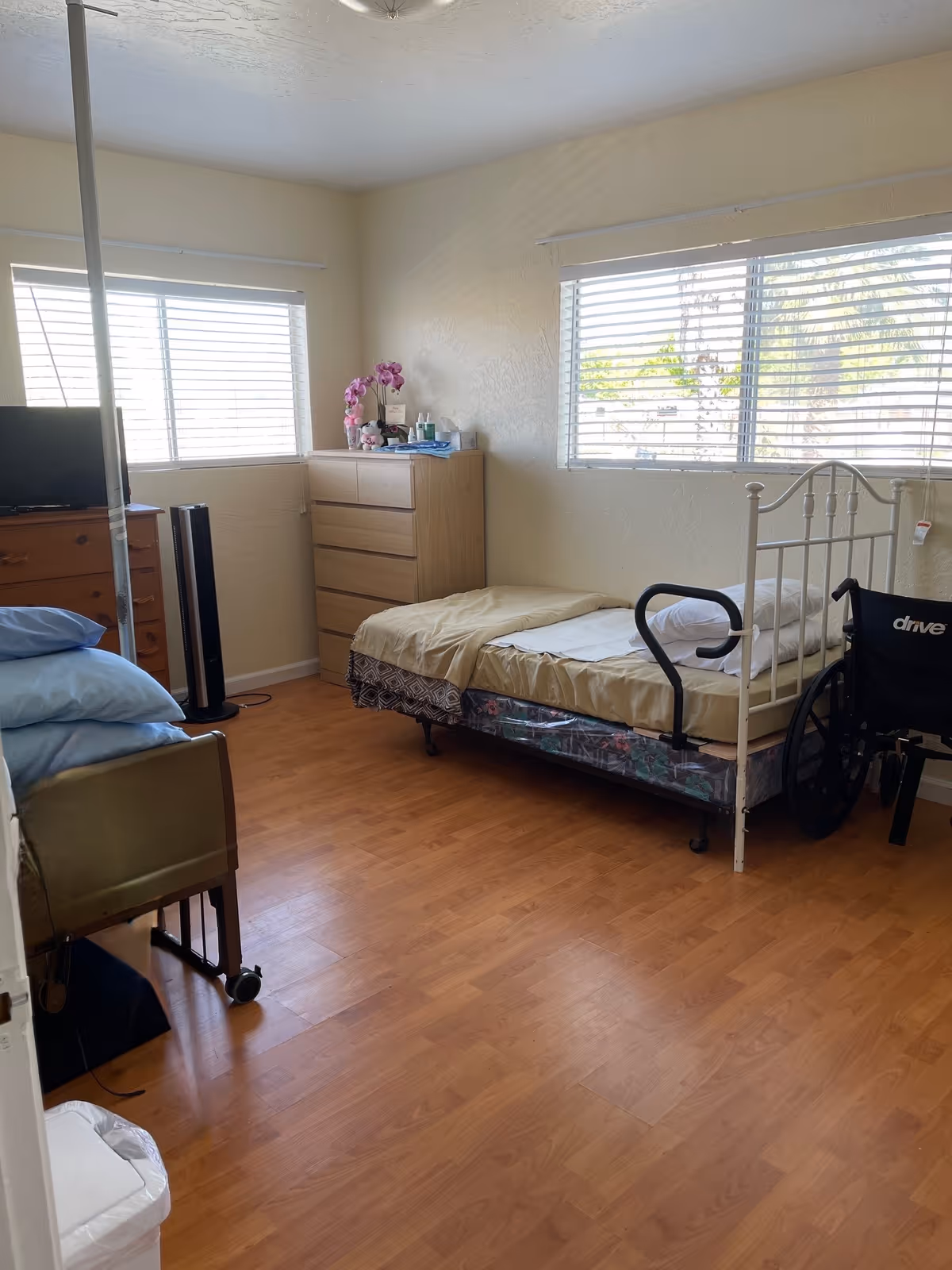 Well-lit resident bedroom with a single metal bed, wheelchair, dresser, TV and wood laminate floor.