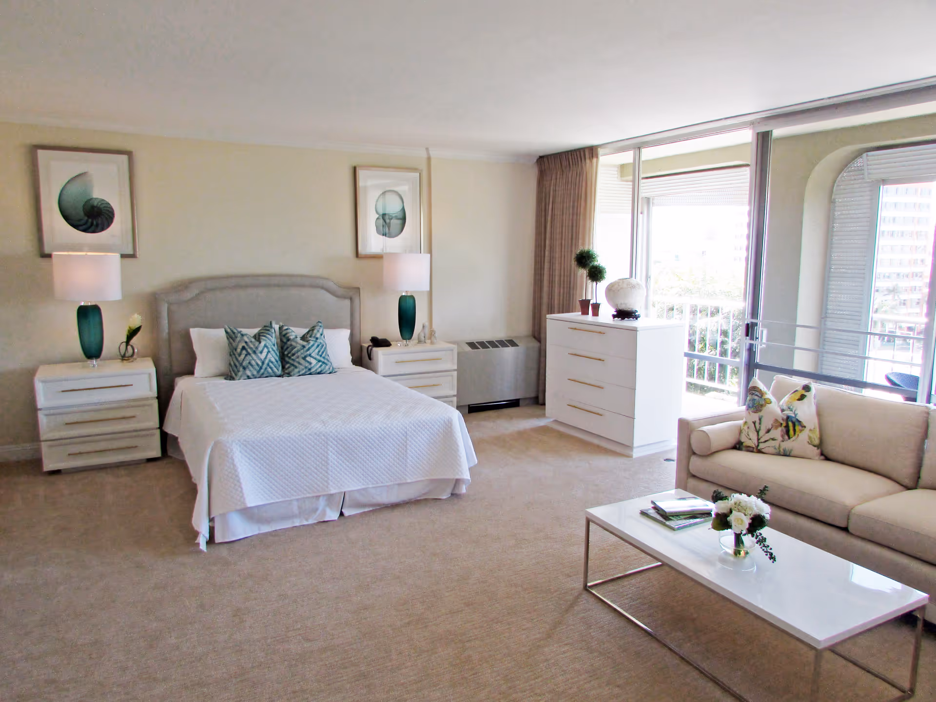 A bright and spacious bedroom with a large bed centered between two nightstands, each with a lamp and decorative items. The bed has white bedding and three patterned pillows. Above the bed are two framed shell prints. To the right, there is a white dresser with decorative plants and a large window with sliding glass doors leading to a balcony. In front of the window is a beige sofa with two floral pillows and a white coffee table with a small flower arrangement and books.