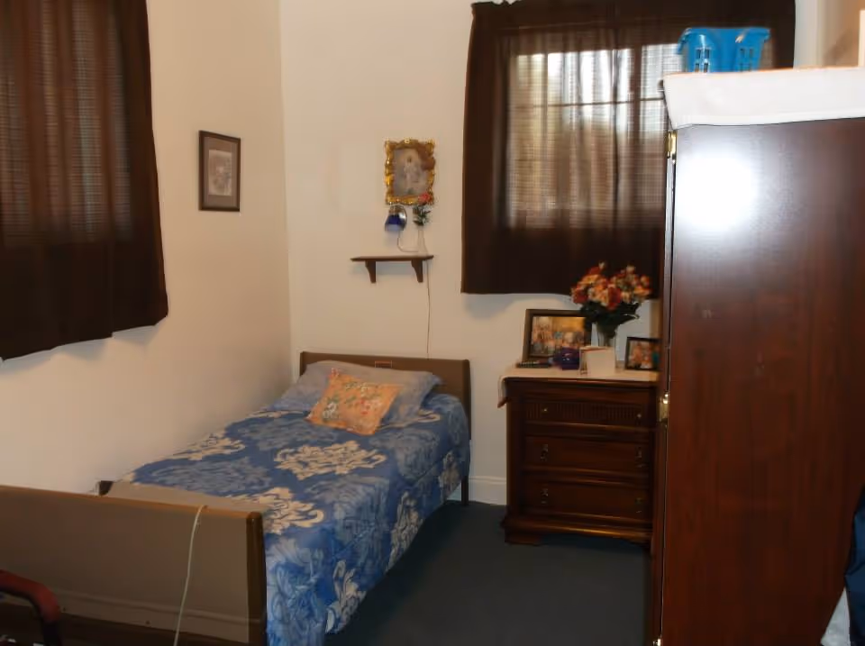 A small bedroom with a single bed covered in a blue floral bedspread and a decorative pillow. There are two windows with dark curtains, a wooden dresser with framed photos and a vase of flowers on top, and a wooden wardrobe. A small shelf on the wall holds a framed picture and a small vase with flowers.