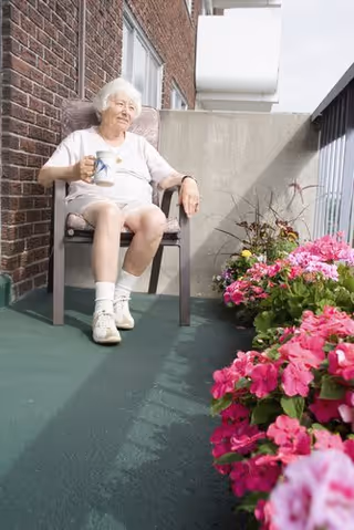 An elderly woman with white hair sitting on a chair on a balcony or patio, holding a cup. She is wearing a white outfit and white shoes. There are vibrant pink flowers in the foreground along the edge of the balcony, and a brick wall and railing are visible in the background.