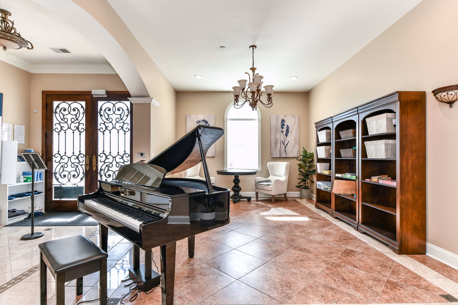 Sunlit common room with a black grand piano, piano bench, bookshelves, and seating near decorative double doors.