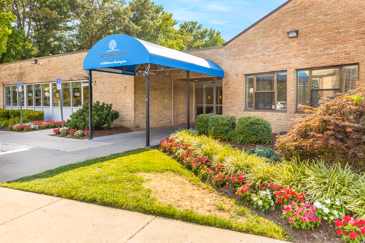 Entrance of Autumn Lake Healthcare with a blue awning over the doorway, brick facade, and landscaped flower beds.