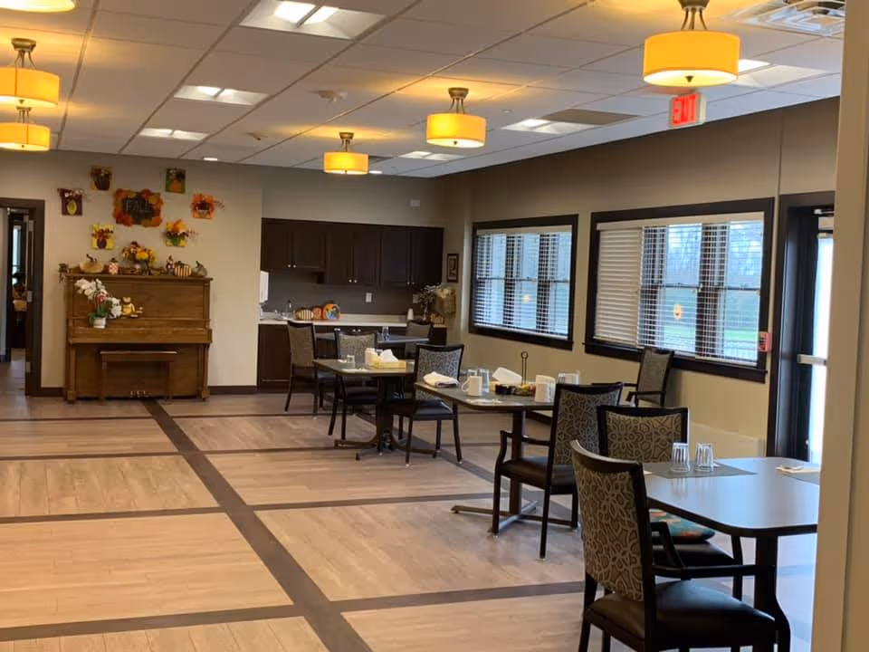 Interior view of a dining area in an assisted living facility with several tables and chairs arranged neatly. The room has large windows with blinds, a piano against the wall decorated with fall-themed items, and ceiling lights providing warm illumination. There is a small kitchenette area with cabinets and a counter in the background.