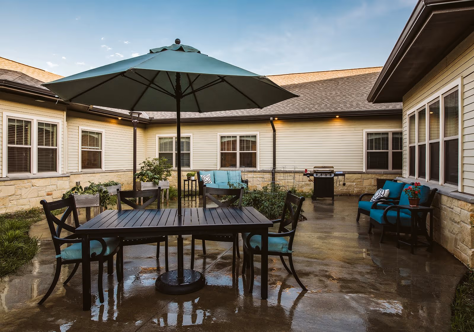 Outdoor courtyard patio with a table, umbrella, chairs, and cushioned seating beside a single-story building.