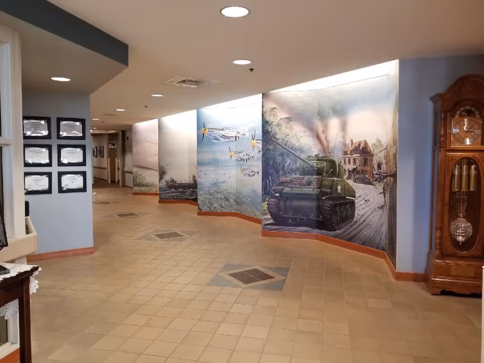 Interior hallway of Norfolk Veterans' Home with a mural depicting military scenes including a tank, airplanes, and a naval ship. The hallway has tiled flooring, recessed ceiling lights, framed certificates on the left wall, and a large wooden grandfather clock on the right.