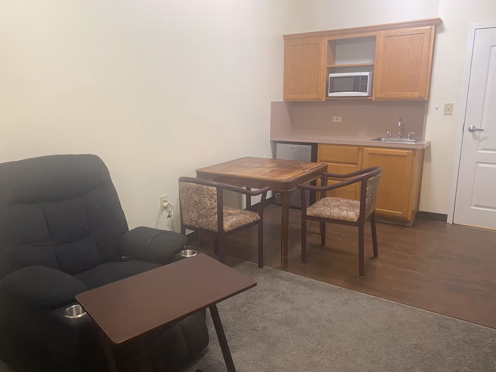 A small living area with a black recliner chair equipped with cup holders and a small brown table in front of it. Behind the chair is a wooden dining table with two floral-patterned chairs. On the back wall, there is a kitchenette with wooden cabinets, a microwave, a small refrigerator, and a sink. The floor is a combination of carpet and wood.