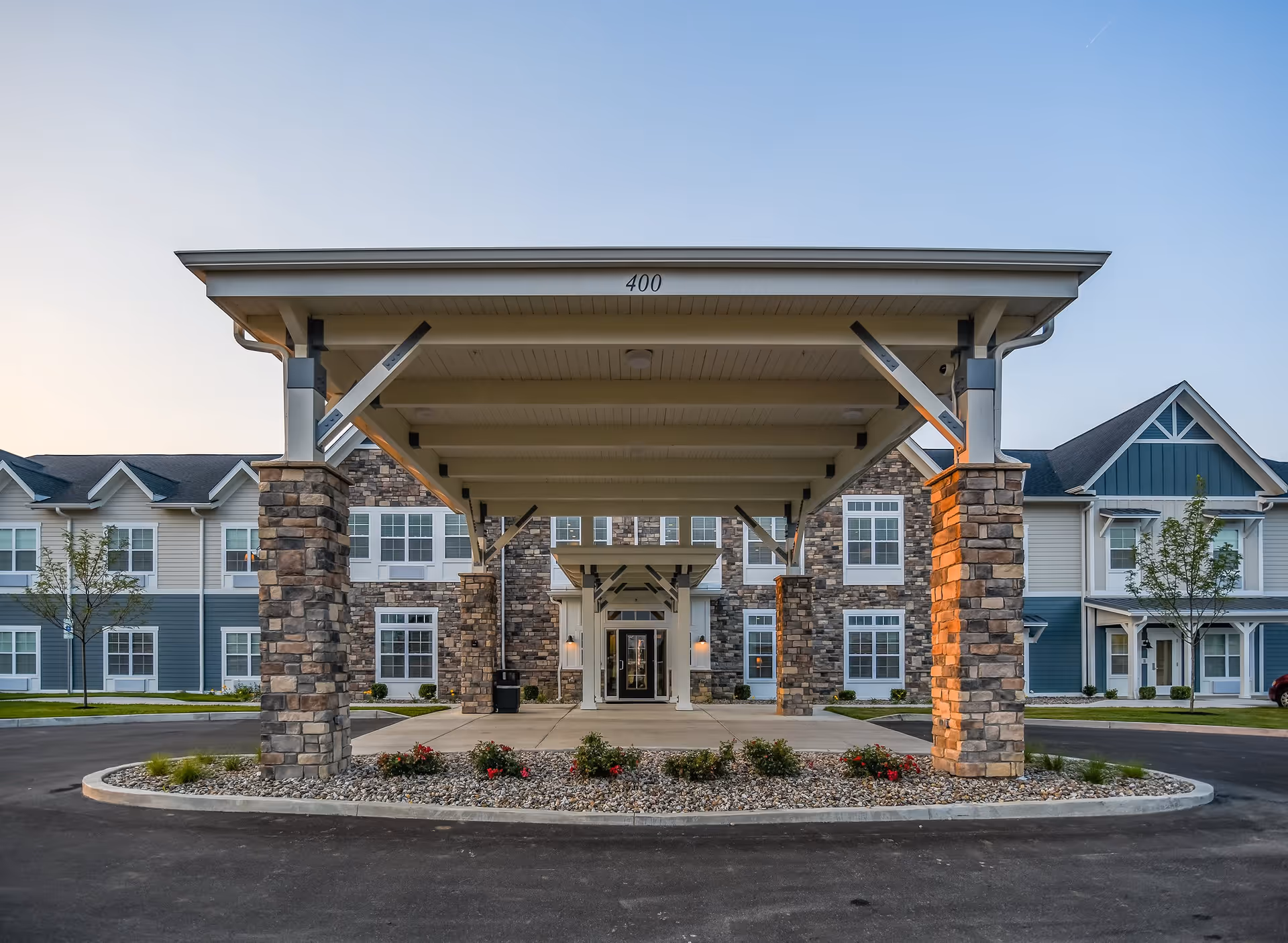 Front entrance of a senior living facility with a covered drop-off area supported by stone pillars, surrounded by a landscaped area with small bushes and flowers, and a two-story building with blue and beige siding in the background under a clear sky.