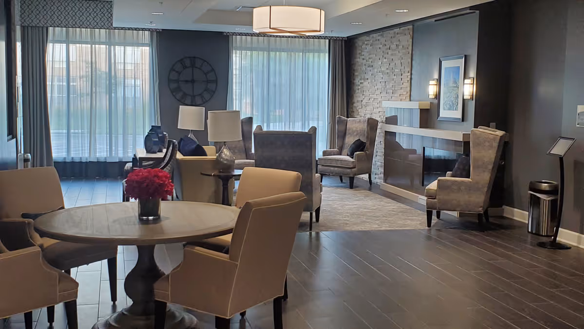 A cozy senior living common area with a round wooden table and four beige chairs in the foreground, a vase with red flowers on the table, and several armchairs arranged around a modern fireplace with a stone accent wall. Large windows with sheer curtains allow natural light to fill the room.