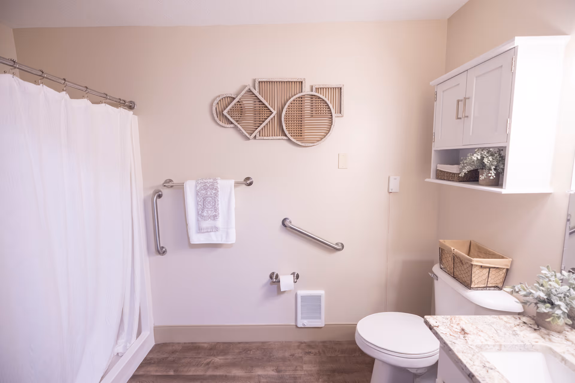 A clean and well-lit bathroom with a white shower curtain on the left, grab bars on the wall, a towel hanging on one of the bars, a toilet with a basket on top, a white cabinet mounted on the wall above the toilet, and decorative wall art above the towel rack. The floor has a wood-like finish and there is a small heater vent near the baseboard.