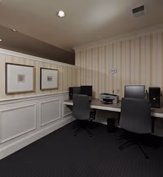 A small office area with two black office chairs in front of a desk that holds two desktop computers and a printer. The room has striped wallpaper and white wainscoting, with two framed pictures on the wall and a carpeted floor.