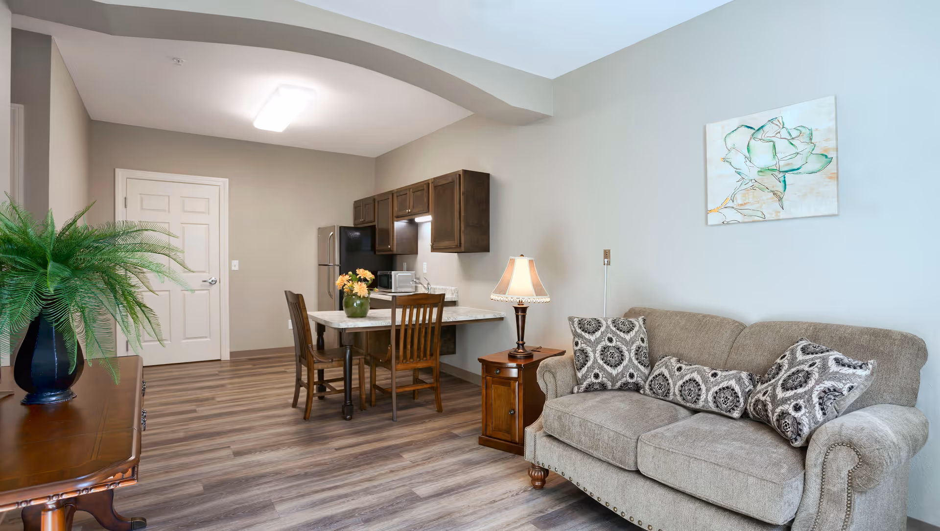 A cozy living area with a beige sofa adorned with patterned cushions, a wooden side table with a lamp, and a small dining table with two chairs. The background features a compact kitchen with dark wooden cabinets, a refrigerator, and a microwave. A green potted plant sits on a wooden table to the left, and a framed artwork of a flower hangs on the wall above the sofa.
