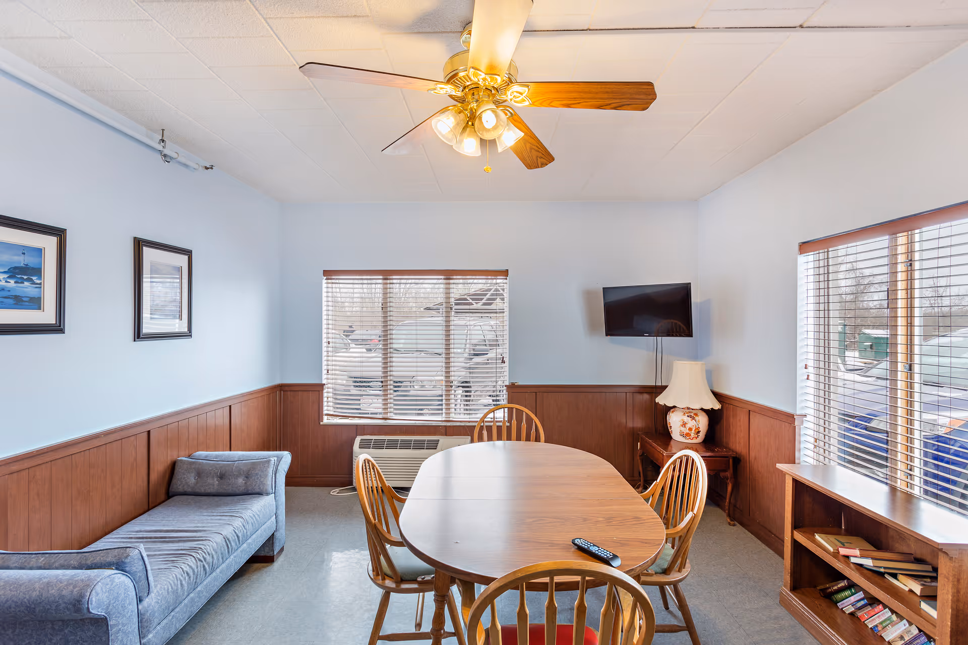 A communal interior room with a wooden dining table and chairs, a chaise lounge, ceiling fan, wall-mounted TV, and windows with blinds.