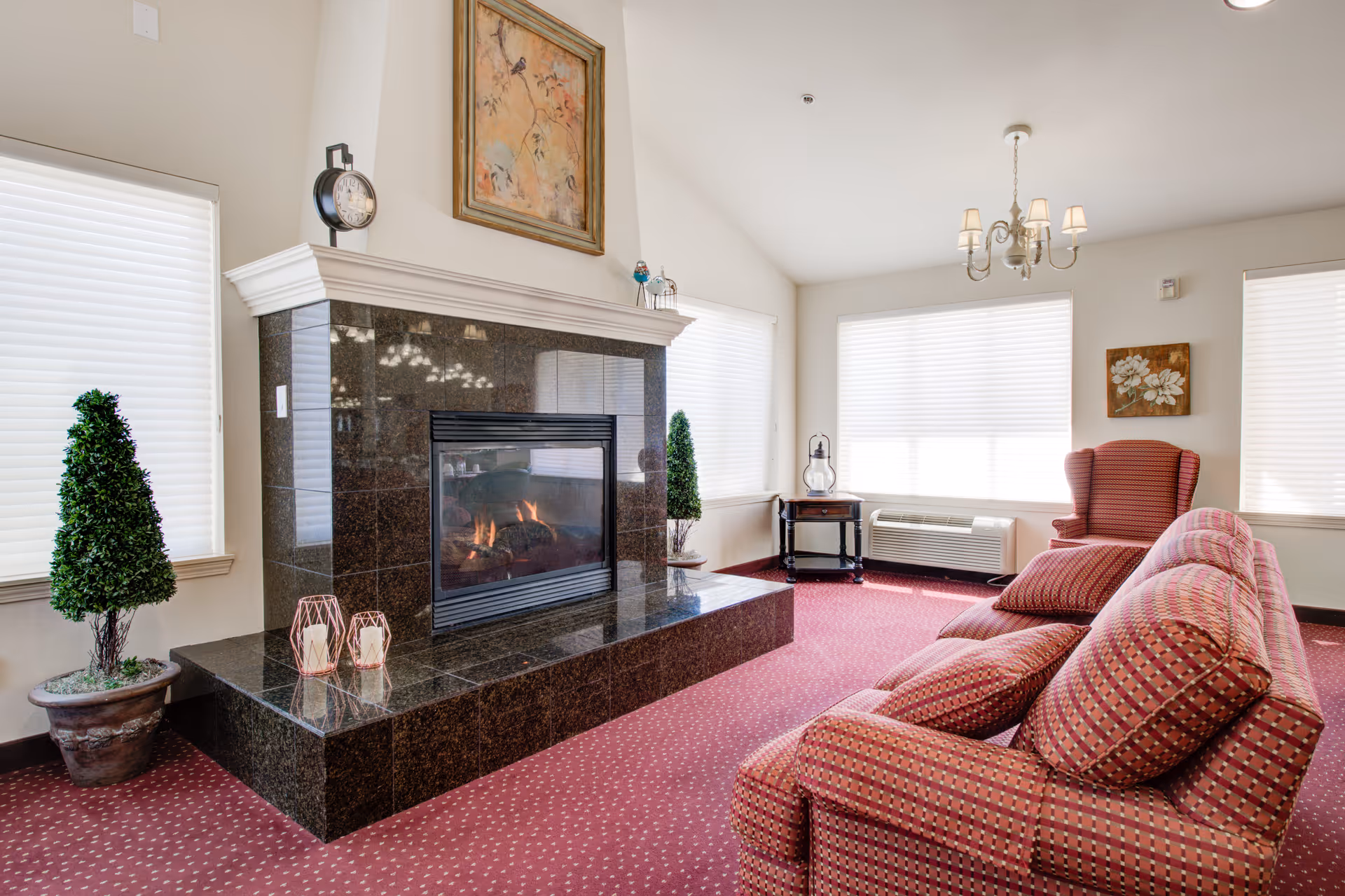 Sunlit senior living room with a marble fireplace, red patterned sofas and armchair, potted topiaries, and large windows.