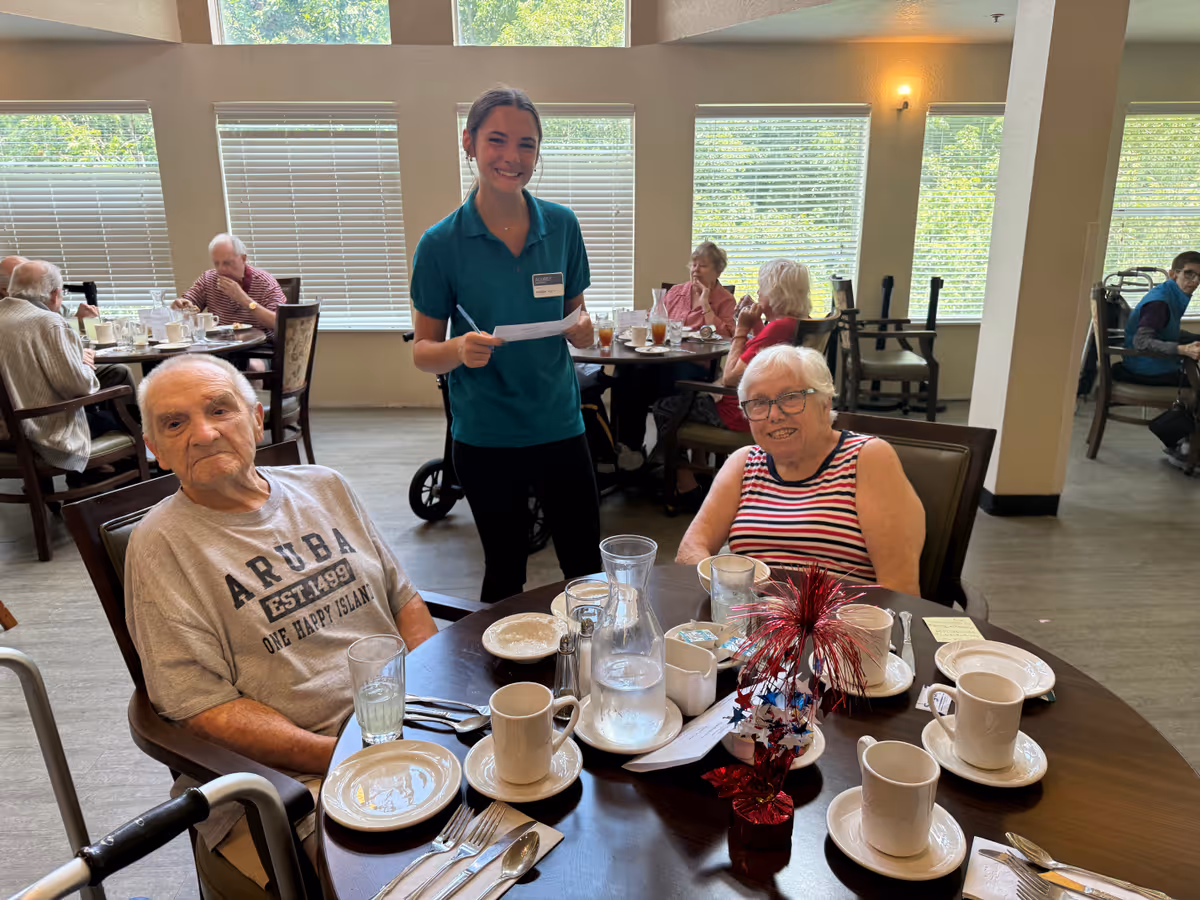 A dining room in a senior living facility with elderly residents seated at tables enjoying a meal. A smiling staff member in a teal shirt stands next to a table with two elderly individuals, one man wearing an Aruba t-shirt and one woman in a striped sleeveless top. The table has cups, plates, utensils, and a patriotic red, white, and blue centerpiece. Large windows with blinds allow natural light into the room.