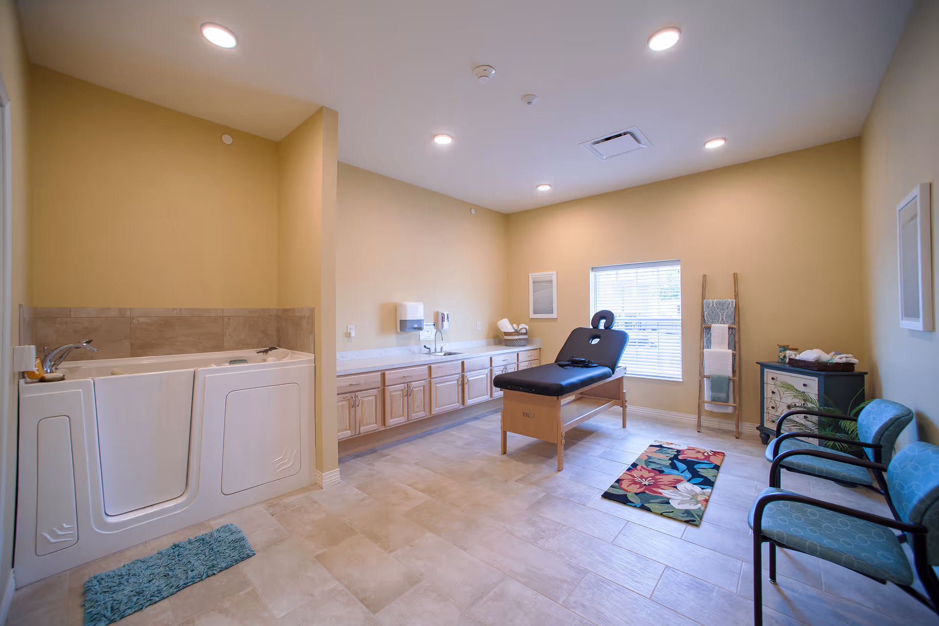 A spacious therapy room with a walk-in bathtub on the left, a black massage table in the center, a long countertop with cabinets and a sink along the back wall, two blue chairs on the right, a colorful floral rug on the floor, and a window with blinds letting in natural light.