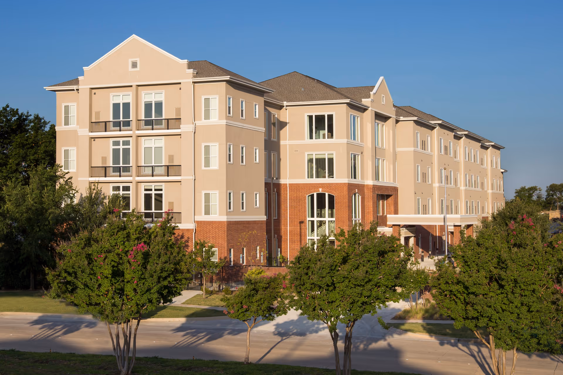 Exterior view of a multi-story senior living facility building with beige and brick facade, surrounded by green trees and a clear blue sky.