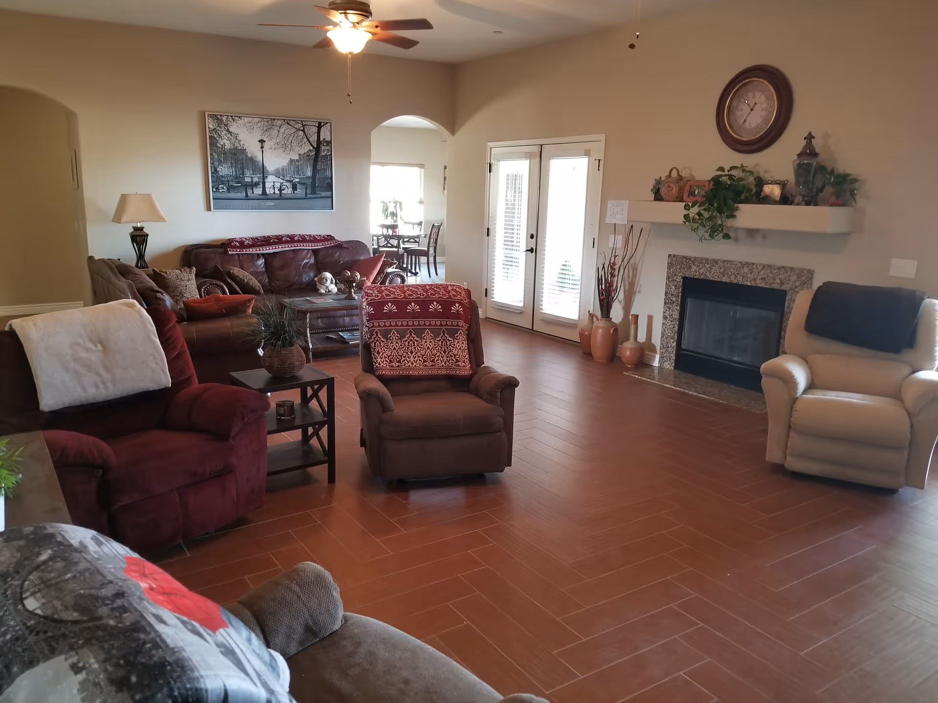 A cozy living room with multiple armchairs and a sofa arranged around a coffee table. The room features a ceiling fan with a light, a fireplace with decorative items and plants on the mantel, and a large wall clock above it. French doors with blinds lead to another area, and a black and white framed photo hangs on the wall above the sofa.