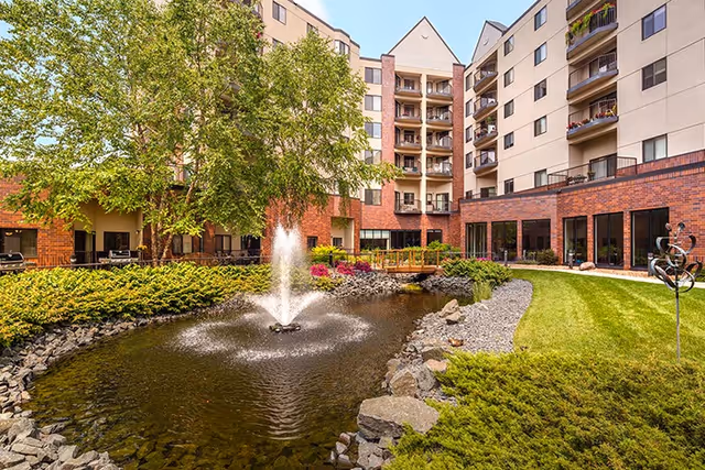 Outdoor courtyard area of a senior living facility with a pond featuring a central water fountain, surrounded by rocks, greenery, and trees. The multi-story building with balconies and large windows encloses the courtyard.