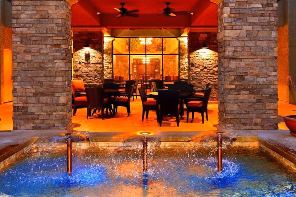 Outdoor seating area at night with stone pillars and a water fountain in the foreground. The seating area has tables and chairs with red cushions, ceiling fans, and warm lighting illuminating the stone walls and interior space visible through large windows.