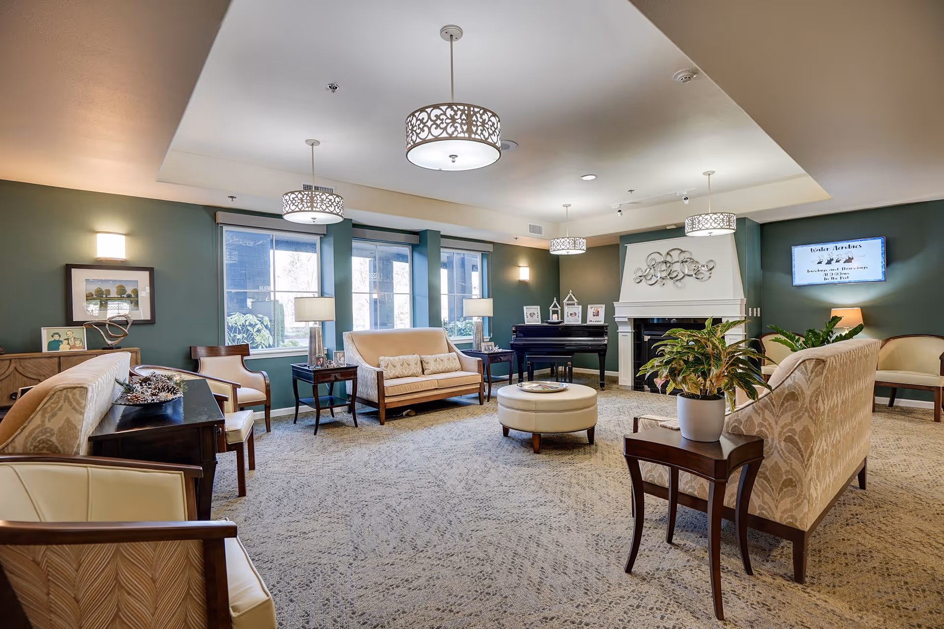 A spacious and well-lit living room in a senior living facility with multiple beige upholstered sofas and chairs arranged around a central round ottoman. The room features green walls, large windows letting in natural light, a white fireplace with decorative metal art above it, a black piano, and several table lamps. There are framed pictures and plants on side tables, and elegant ceiling light fixtures hang from the ceiling.