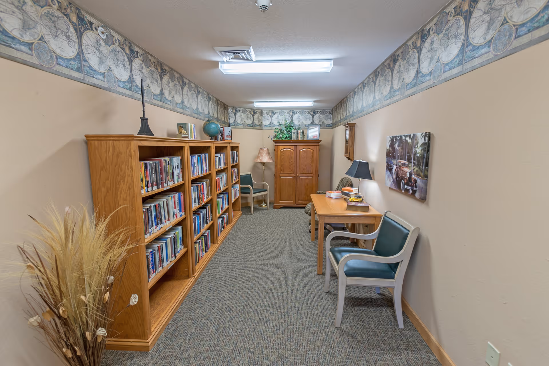 A narrow room with beige walls and carpeted floor featuring two wooden bookshelves filled with books on the left side. At the far end, there is a wooden cabinet and a chair with a floor lamp beside it. On the right side, there is a wooden table with two chairs, a lamp, and some books. The walls have a decorative border near the ceiling and a framed picture of a vintage car on the right wall. A bundle of dried decorative plants is placed in the front left corner.