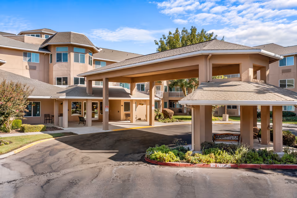 Exterior view of The Hampshire - A Provincial Senior Living Community showing the main entrance with a covered drop-off area, beige building facade, multiple windows, landscaped greenery, and a clear blue sky.