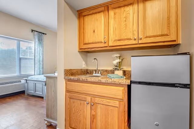 A small kitchenette area with wooden cabinets, a countertop with a sink, a small refrigerator, and a few decorative items including a vase with flowers and folded towels. In the background, a bedroom area with a bed, window with blinds, and curtains is partially visible.