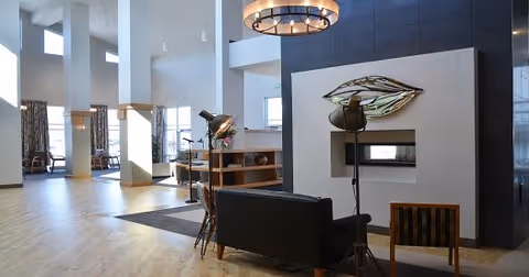 A spacious and modern common area with high ceilings and large windows allowing natural light to fill the room. The space features a contemporary fireplace with artistic metal wall decor above it, a black sofa, a wooden chair, and a small table with a lamp. Light wood flooring and neutral-colored walls create a warm and inviting atmosphere.