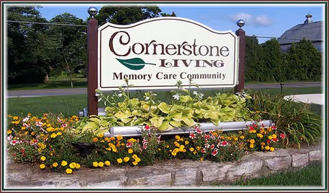 Outdoor sign for Cornerstone Living Memory Care Community surrounded by a flower bed with various colorful flowers and greenery, set on a stone border with grass and trees in the background.
