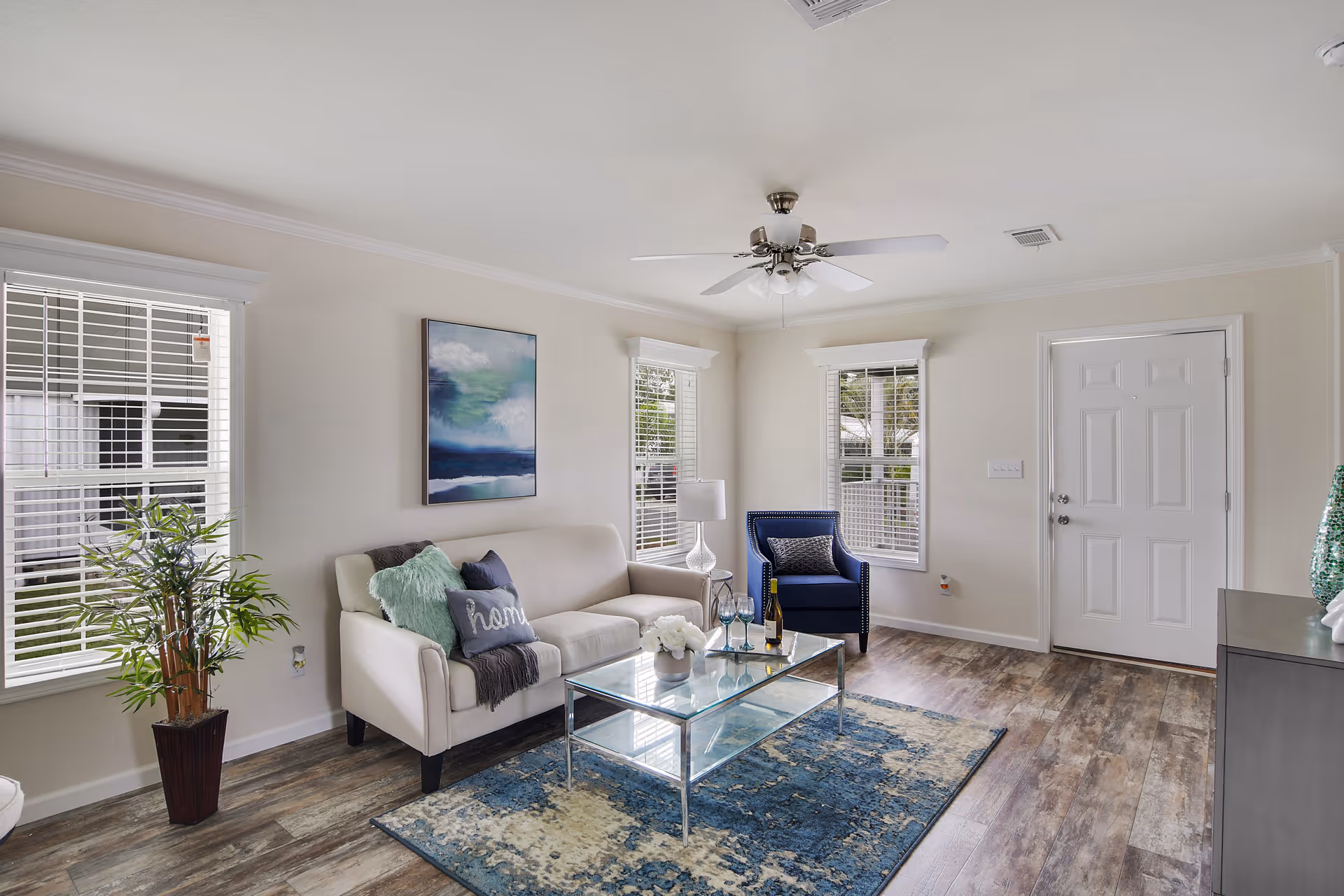 A bright living room with a white sofa adorned with pillows and a throw blanket, a blue armchair, a glass coffee table with decorative items, a blue patterned rug, a ceiling fan, and three windows with white blinds letting in natural light. There is a potted plant near one window and a painting on the wall above the sofa.