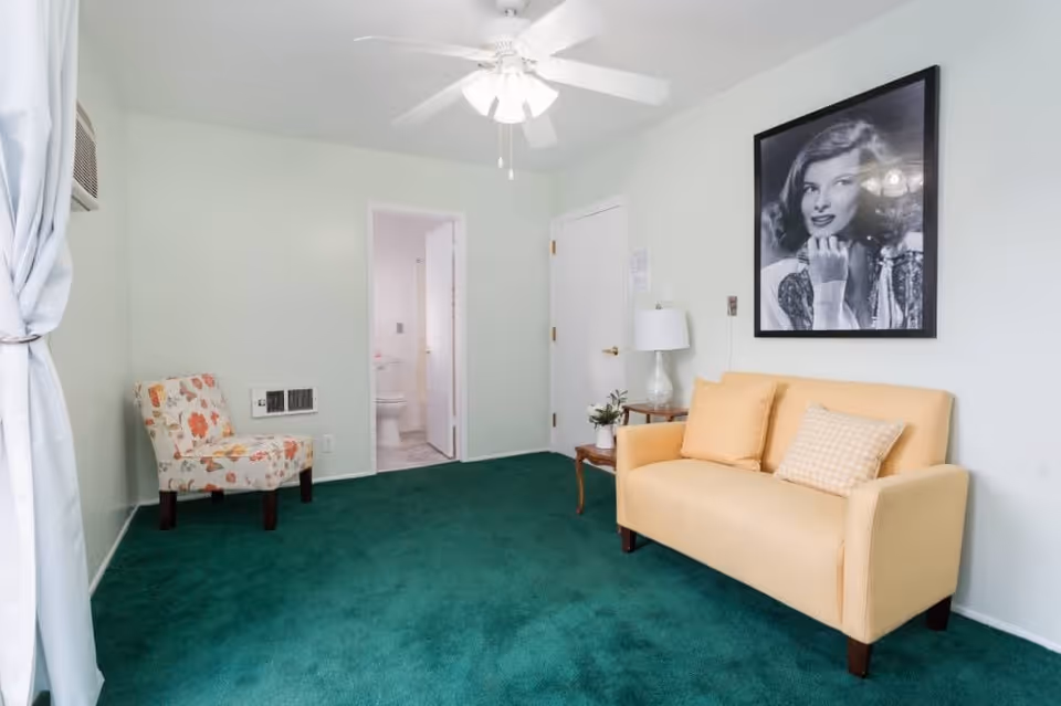 A small living room with green carpet, a yellow loveseat with two pillows, a floral patterned chair, a side table with a lamp and plant, a ceiling fan with light, and a black and white portrait of a woman on the wall. There is a doorway leading to a bathroom with a visible toilet.
