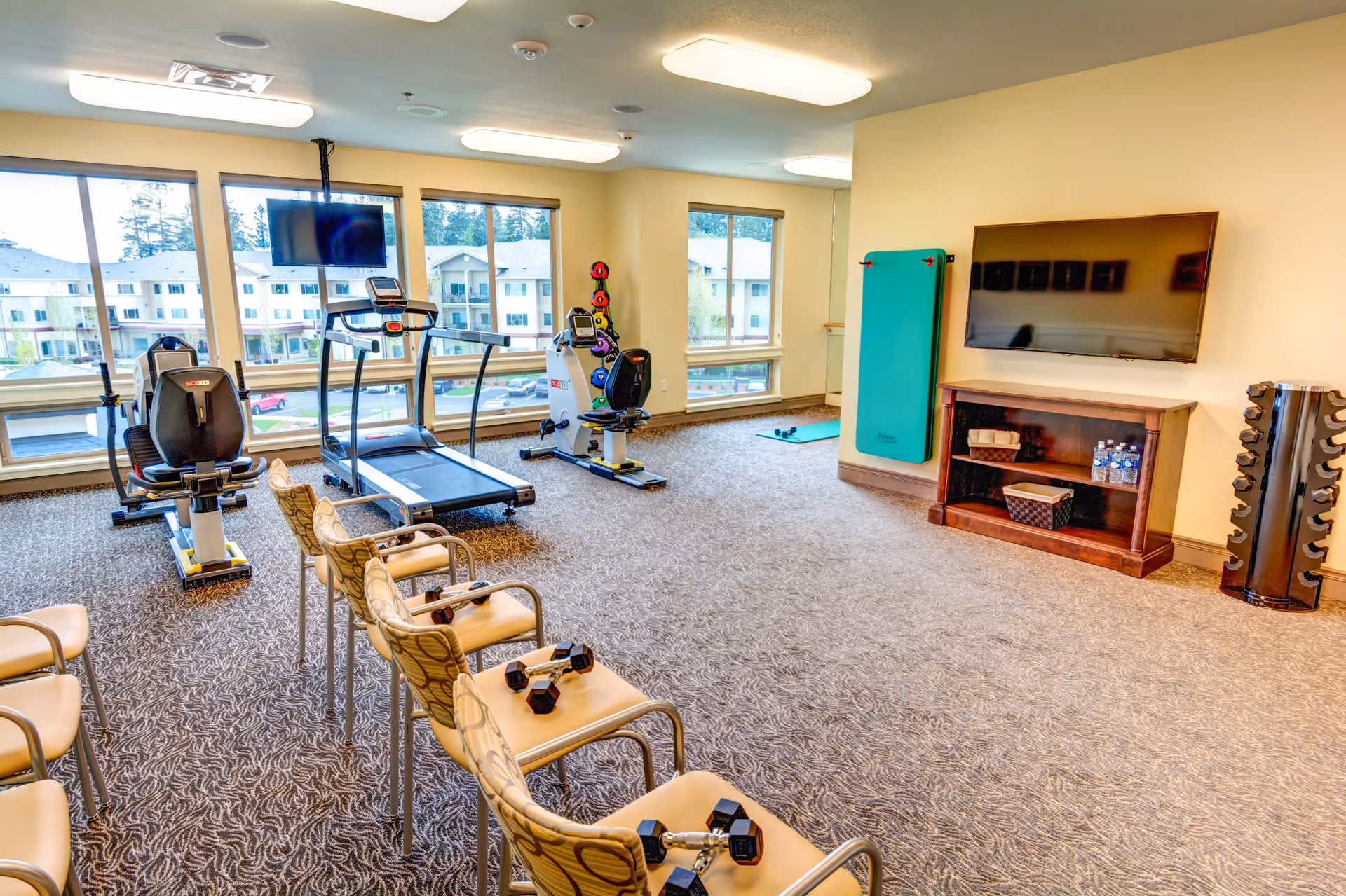 A fitness room in a senior living facility with exercise equipment including a treadmill, stationary bikes, and dumbbells. Several chairs with dumbbells on them are arranged facing a wall-mounted TV. Large windows provide natural light and a view of nearby buildings.