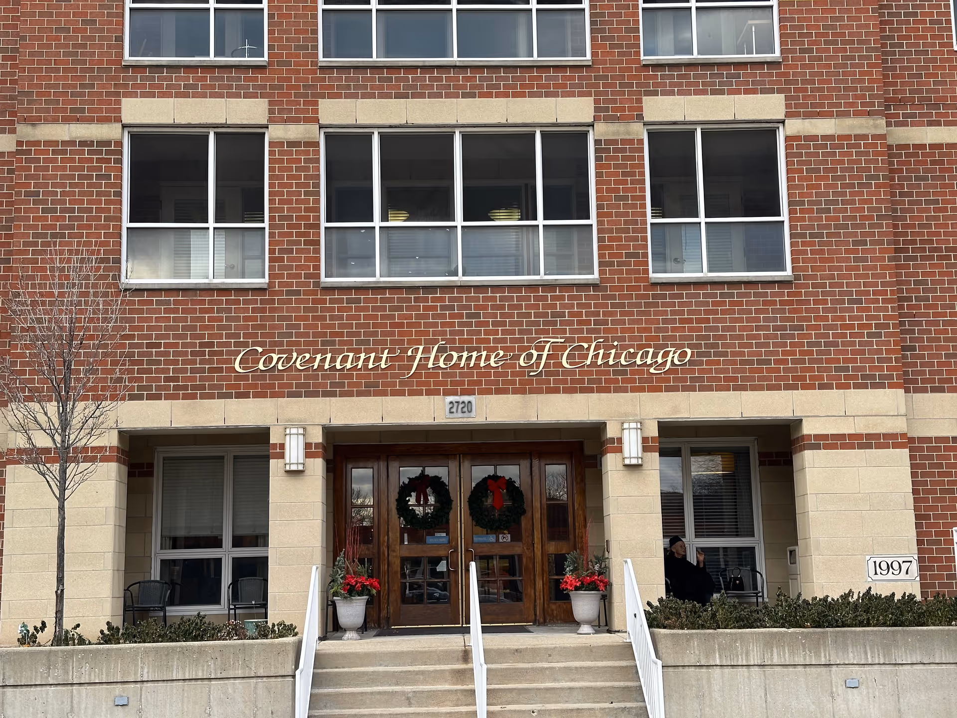 Front entrance of Covenant Home of Chicago, a brick building with large windows. The entrance has double wooden doors decorated with wreaths and flanked by two planters with red flowers. There are stairs leading up to the entrance and a person sitting on a bench to the right side under a covered area.