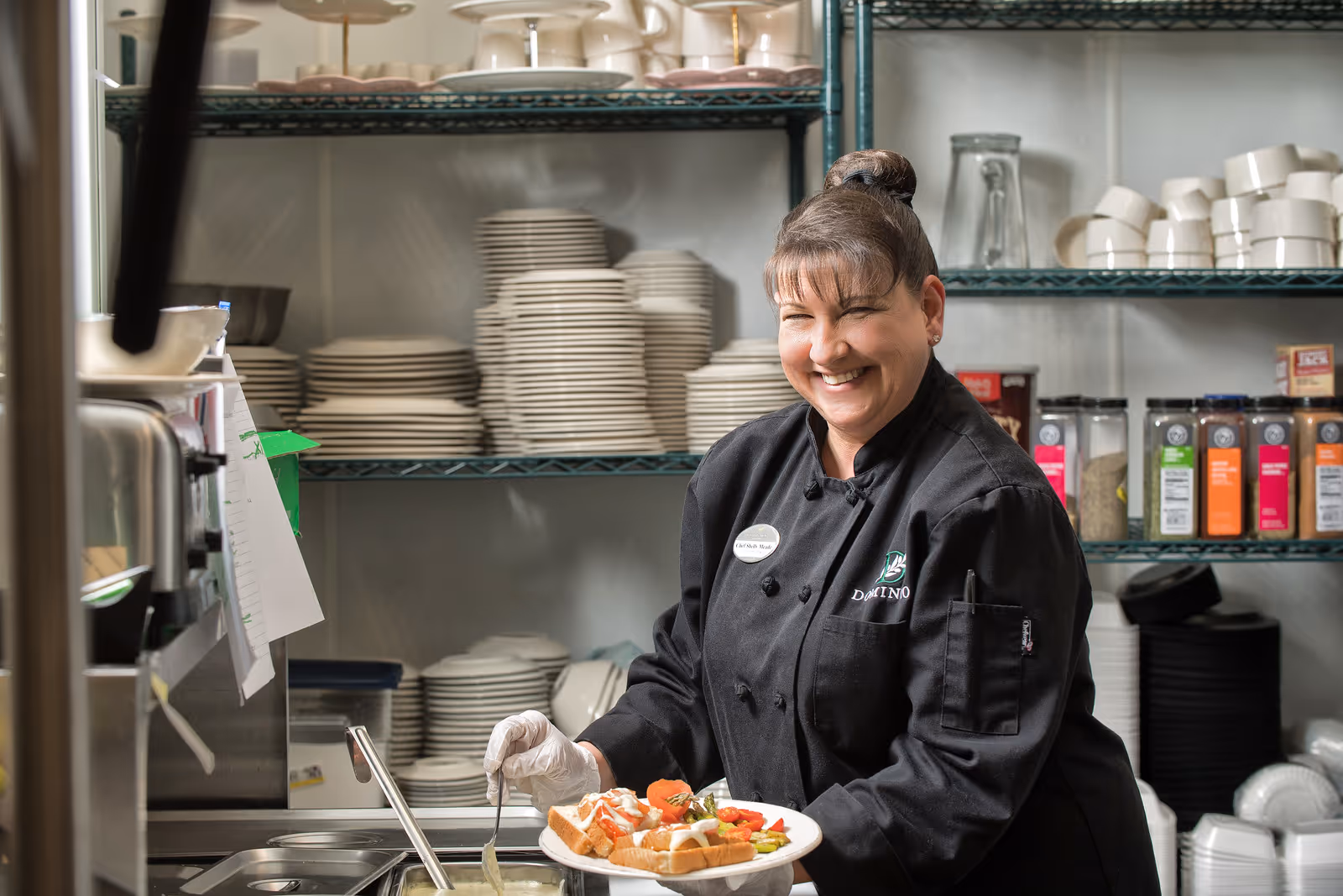 A smiling chef in a black uniform and gloves serving a plate of food in a kitchen with shelves stocked with plates, bowls, and various spices in the background.