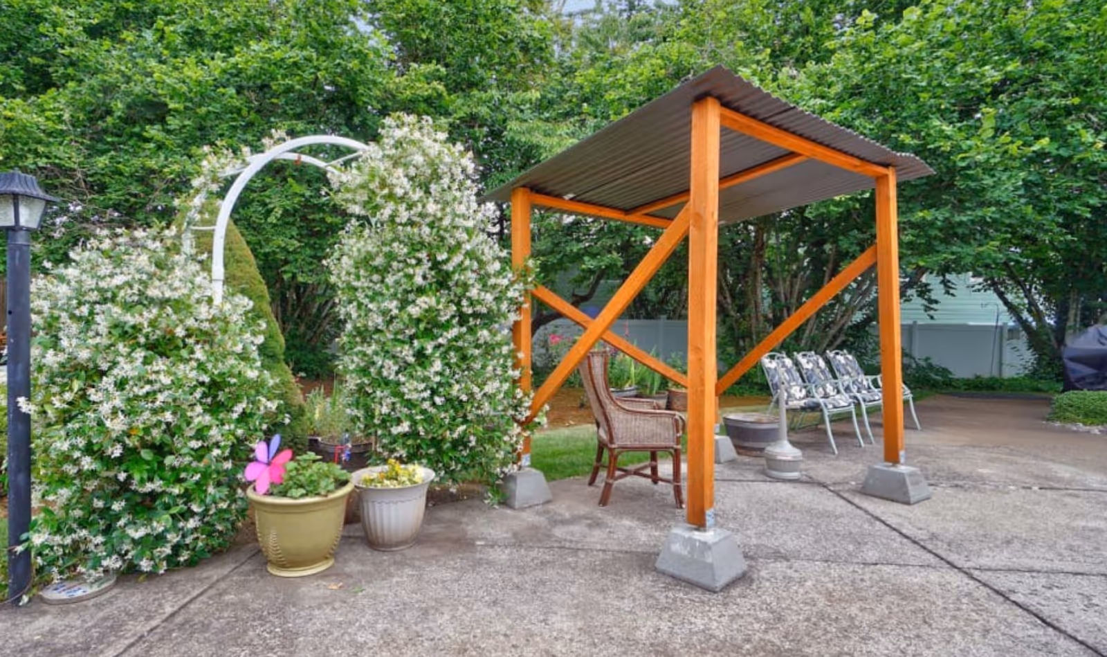 Outdoor patio with a small wooden pergola, seating, potted plants and a flowering vine-covered arch.