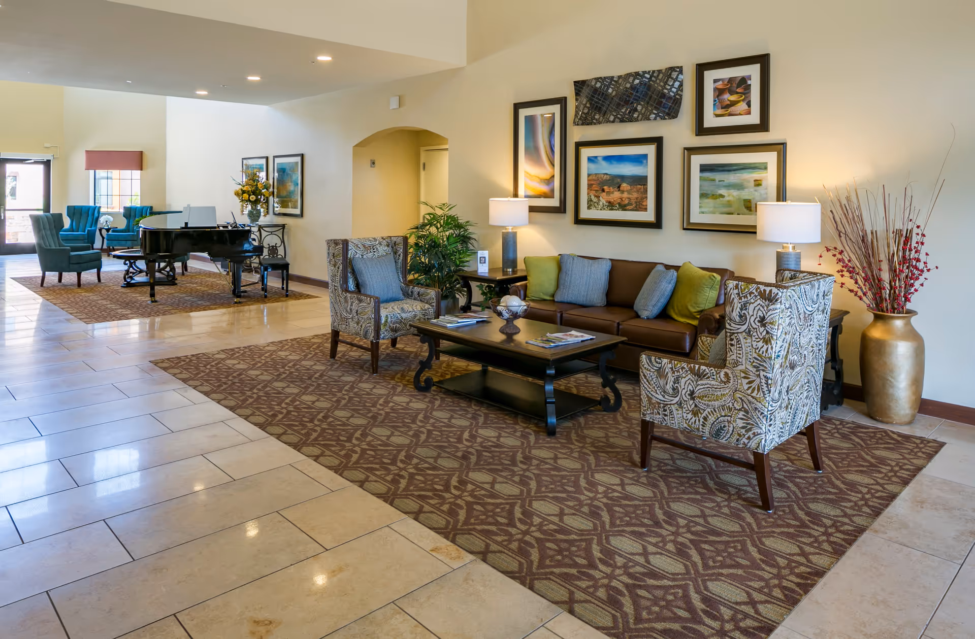 A spacious and well-lit living room area in Orchard Pointe at Arrowhead featuring a brown leather sofa with green and blue cushions, two patterned armchairs, a dark wooden coffee table with magazines, and two table lamps. The walls are decorated with framed artwork. In the background, there is a black grand piano with a chair, two teal armchairs, and a door leading outside. The floor is tiled with beige tiles and partially covered with patterned rugs.