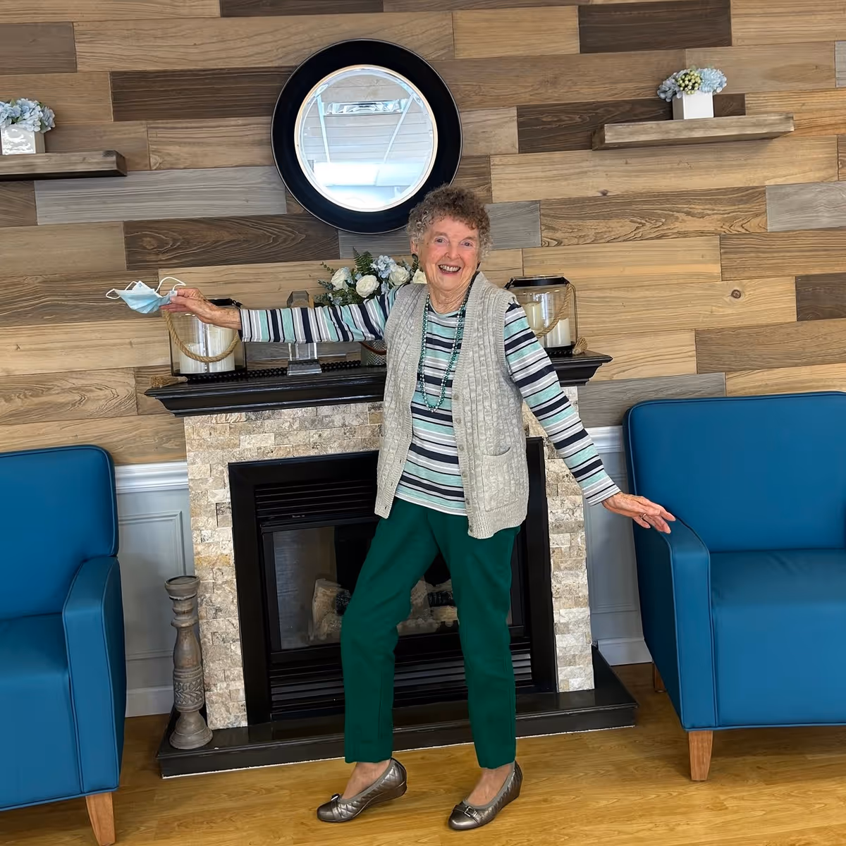 An elderly woman standing and smiling in a cozy room with wooden panel walls, a decorative fireplace, and two blue armchairs. She is holding a face mask in one hand and wearing a striped shirt, green pants, a beige vest, and a beaded necklace. There are small flower arrangements on shelves above the fireplace and a round mirror on the wall.